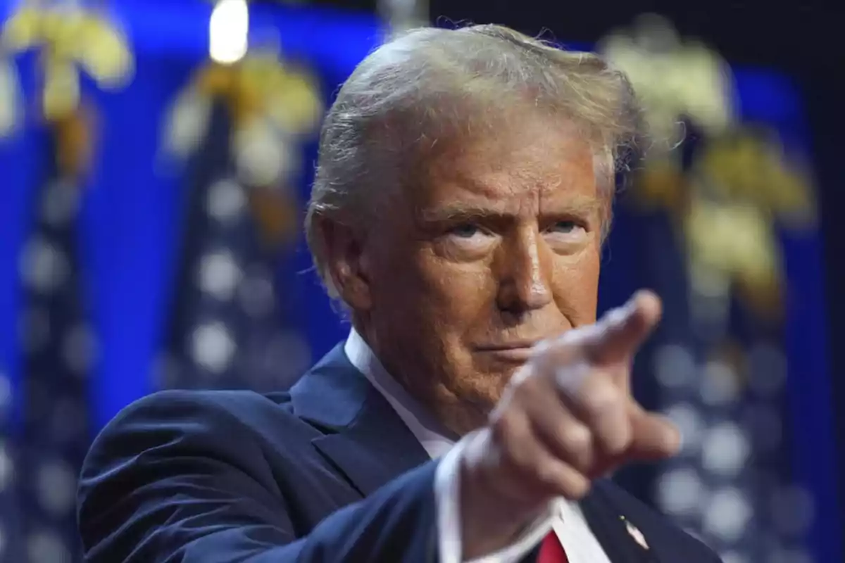 Blond-haired man in a dark suit pointing forward with a serious expression and a background of blurred flags