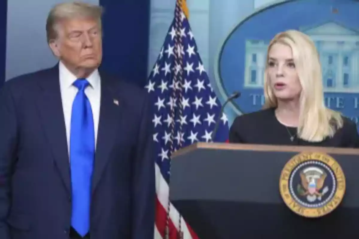 Un hombre con traje azul y corbata azul observa a una mujer rubia que habla en un podio con el escudo presidencial de Estados Unidos y una bandera estadounidense de fondo