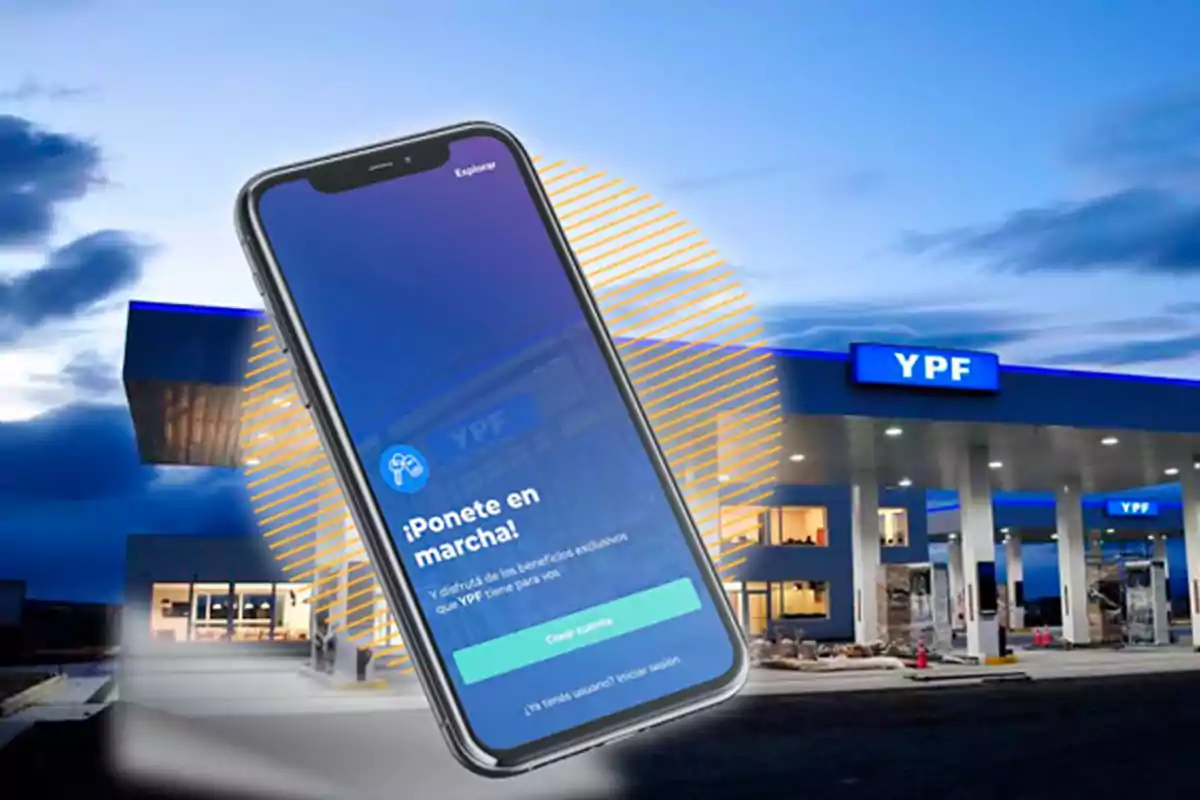 Un teléfono móvil con la pantalla de una aplicación de YPF frente a una estación de servicio YPF al atardecer Un teléfono móvil con la pantalla de una aplicación de YPF frente a una estación de servicio YPF al atardecer