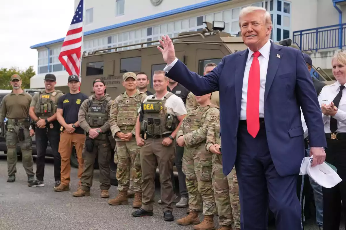 A man in a blue suit and red tie greets in front of a group of agents and uniformed soldiers, with an armored vehicle and a United States flag in the background. A man in a blue suit and red tie greets in front of a group of agents and uniformed soldiers, with an armored vehicle and a United States flag in the background.
