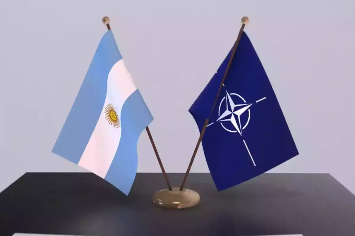Dos banderas pequeñas sobre una mesa, una de Argentina y otra de la OTAN, cruzadas en un soporte dorado Dos banderas pequeñas sobre una mesa, una de Argentina y otra de la OTAN, cruzadas en un soporte dorado