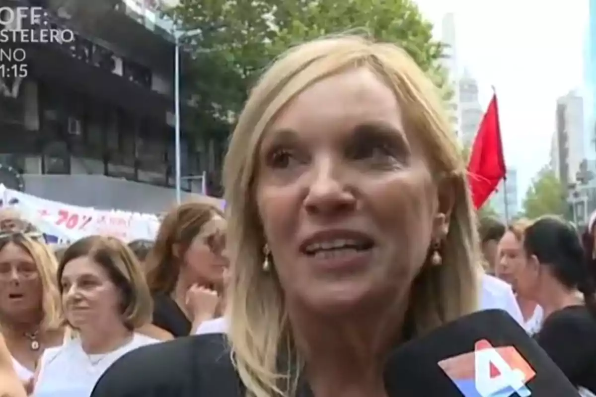 Mujer rubia hablando frente a un micrófono de un canal de televisión durante una manifestación con varias personas y una bandera roja al fondo Mujer rubia hablando frente a un micrófono de un canal de televisión durante una manifestación con varias personas y una bandera roja al fondo