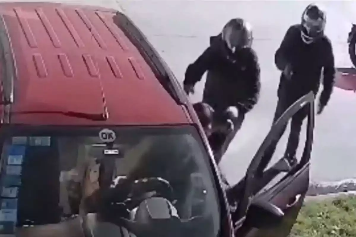 Dos personas con casco se acercan a un auto rojo con la puerta del conductor abierta mientras una persona está sentada al volante Dos personas con casco se acercan a un auto rojo con la puerta del conductor abierta mientras una persona está sentada al volante
