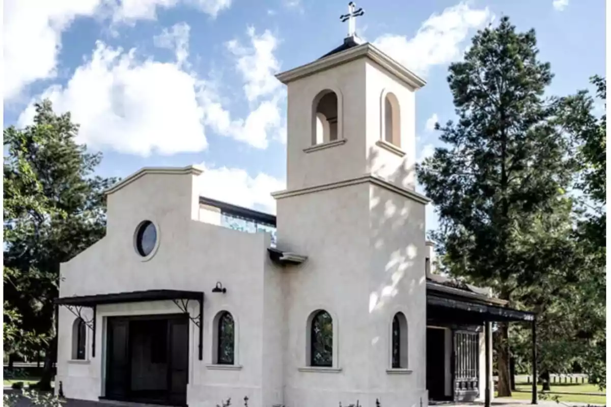 Facade of a small light-colored church with a tower and a cross on top, surrounded by trees and blue sky with clouds Facade of a small light-colored church with a tower and a cross on top, surrounded by trees and blue sky with clouds
