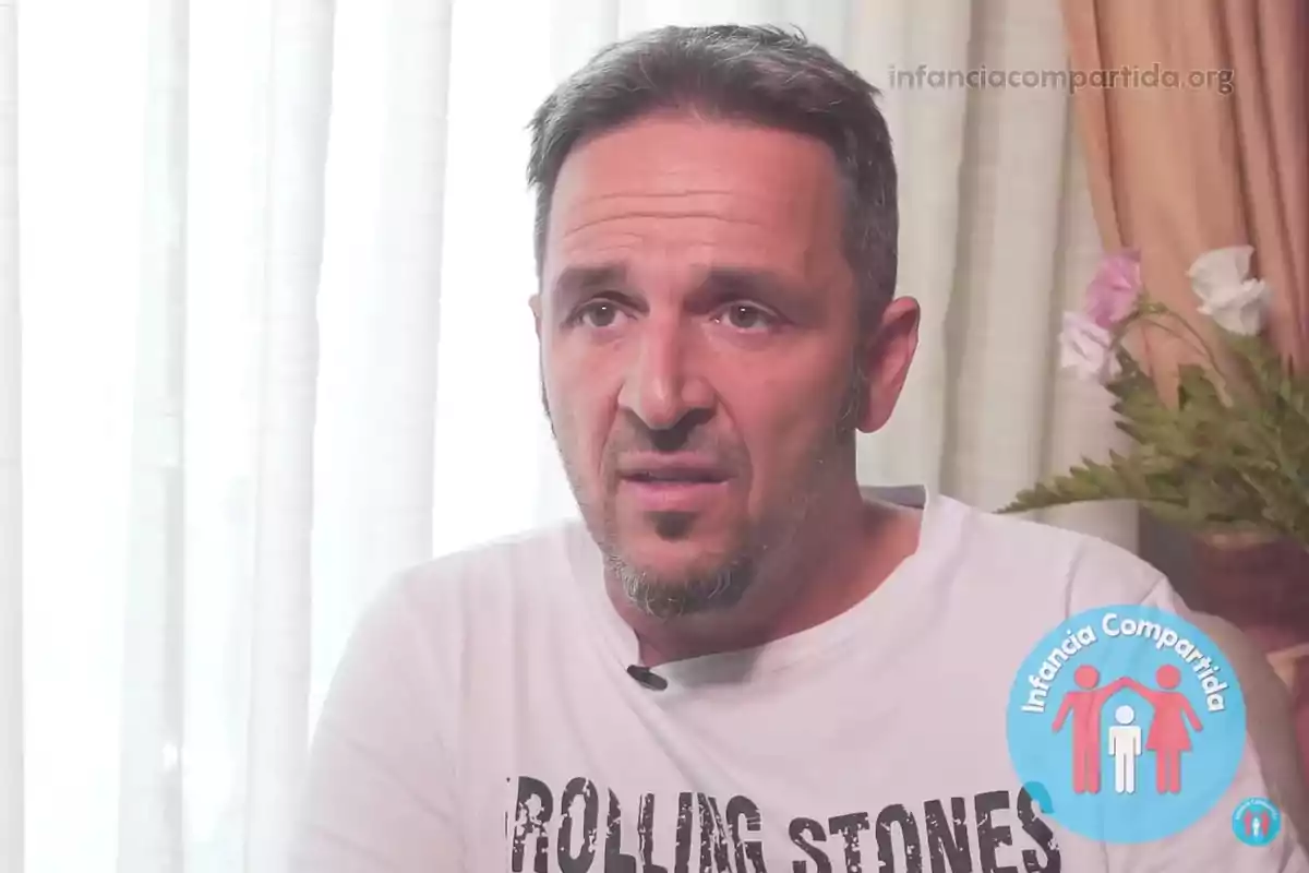 Short-haired man with a trimmed beard wearing a white Rolling Stones t-jersey, sitting in front of a white curtain with flowers in the background and the Infancia Compartida logo in the bottom right corner.