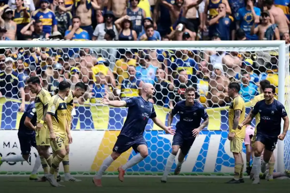 Soccer players celebrate a goal in front of the goal while the crowd in yellow and blue jerseys watches from the stands.