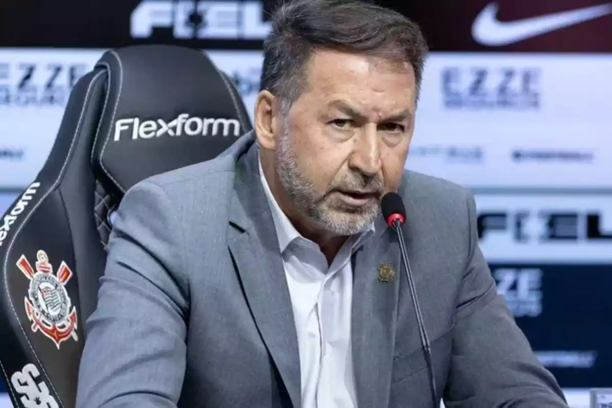 Man in a gray suit speaking at a press conference seated in a chair with the Corinthians club logo Man in a gray suit speaking at a press conference seated in a chair with the Corinthians club logo