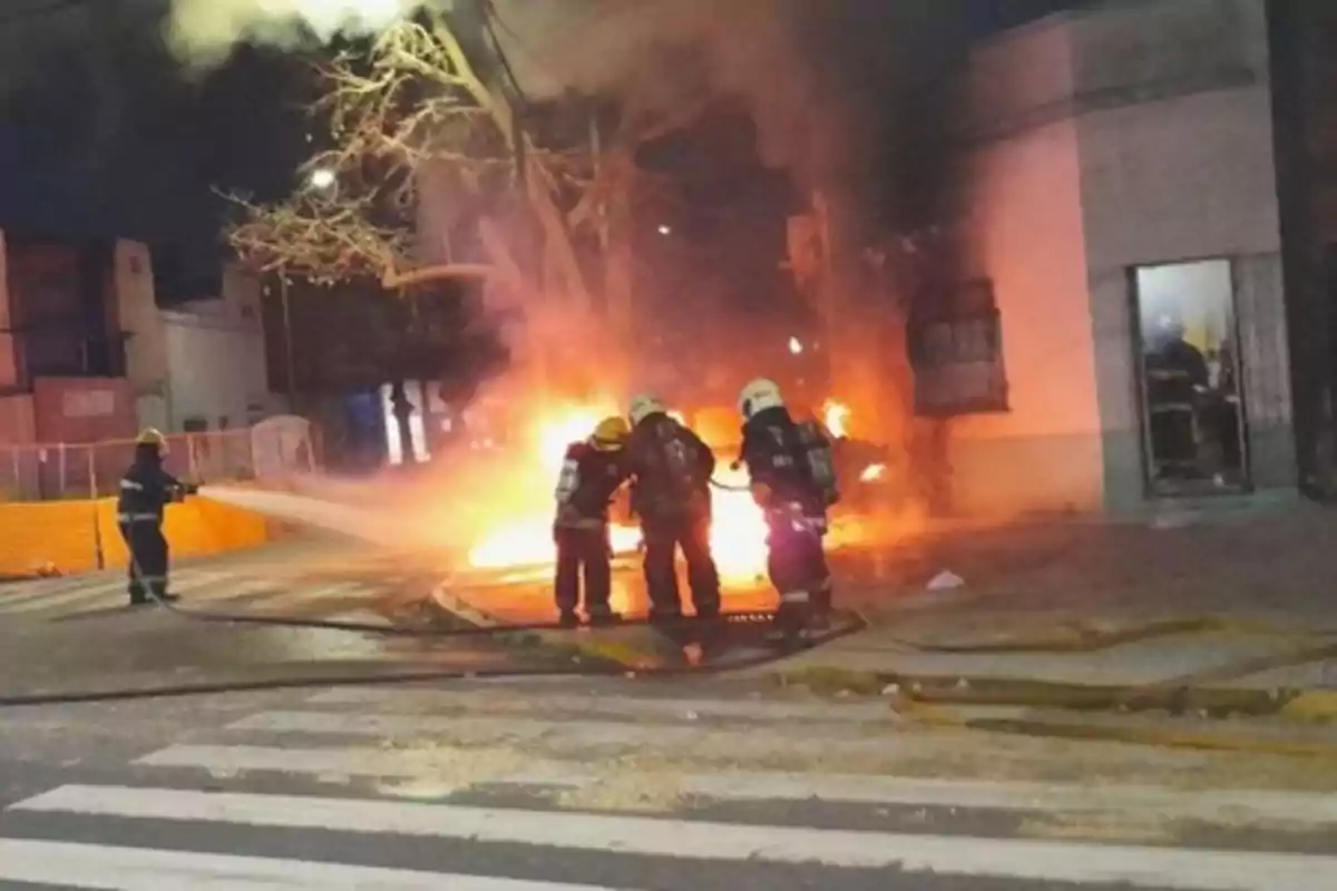Bomberos apagan un incendio en la vía pública durante la noche mientras el fuego y el humo se elevan frente a un edificio Bomberos apagan un incendio en la vía pública durante la noche mientras el fuego y el humo se elevan frente a un edificio