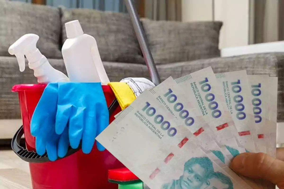Una mano sostiene billetes de diez mil pesos argentinos frente a un balde rojo con guantes azules y productos de limpieza en una sala de estar.