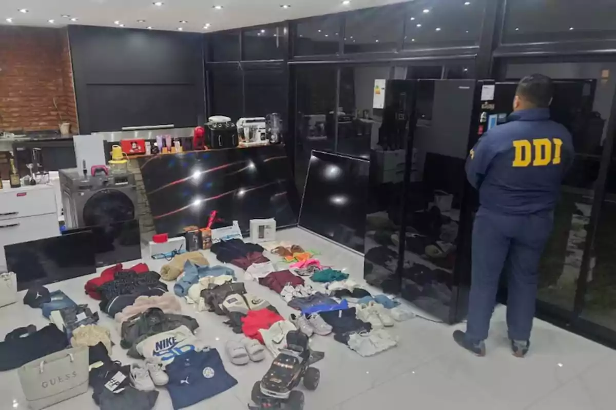 An agent wearing a blue jacket with the initials DDI observes a large amount of clothing, appliances, televisions, and other items organized on the floor of a modern, well-lit room. An agent wearing a blue jacket with the initials DDI observes a large amount of clothing, appliances, televisions, and other items organized on the floor of a modern, well-lit room.