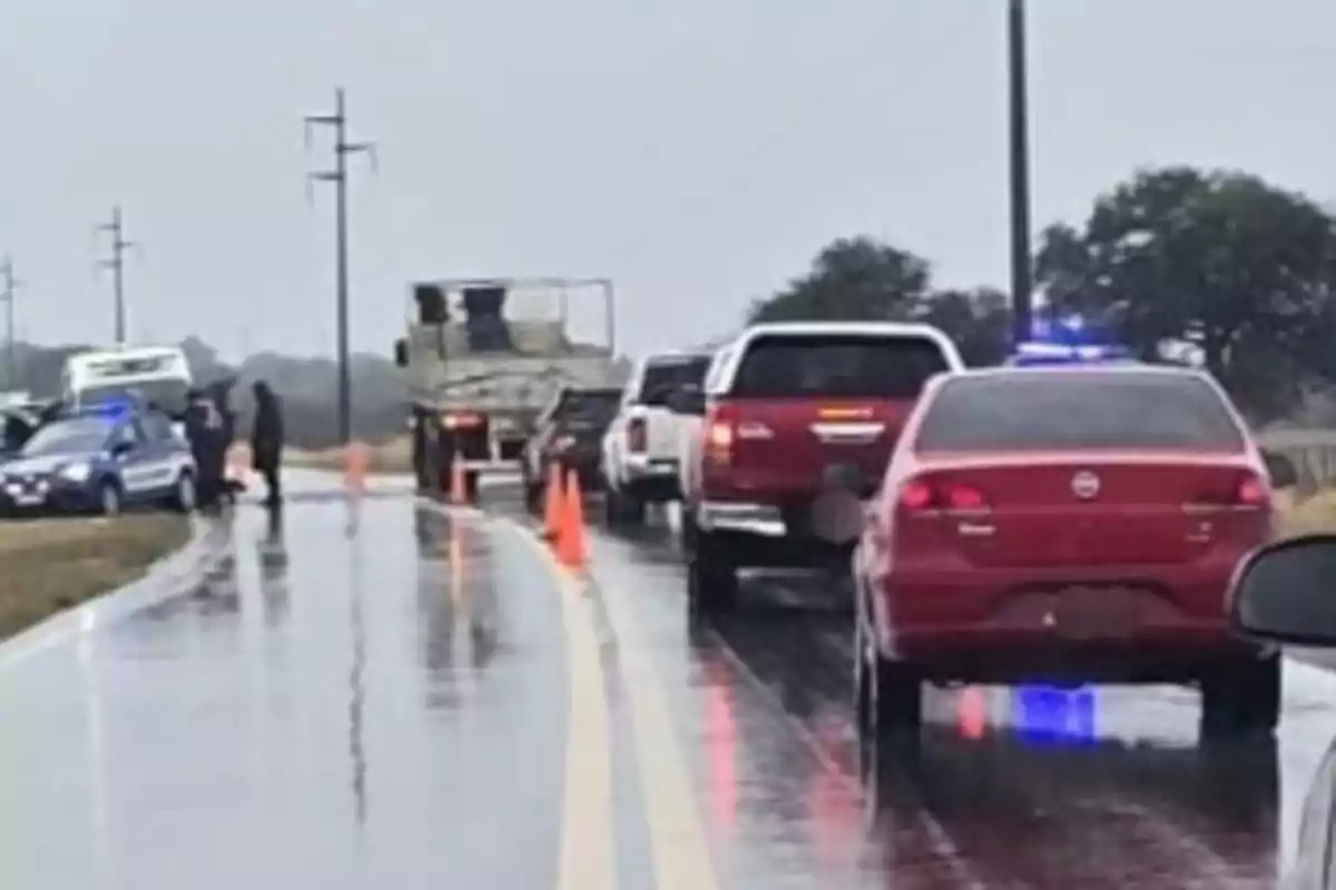 Varios autos y una patrulla están detenidos en una carretera mojada mientras unos policías revisan el área y hay conos naranjas señalando el camino.