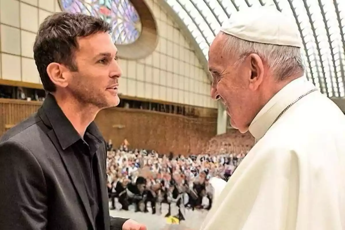 A man in a dark suit talks with the Pope in a room with many people in the background. A man in a dark suit talks with the Pope in a room with many people in the background.
