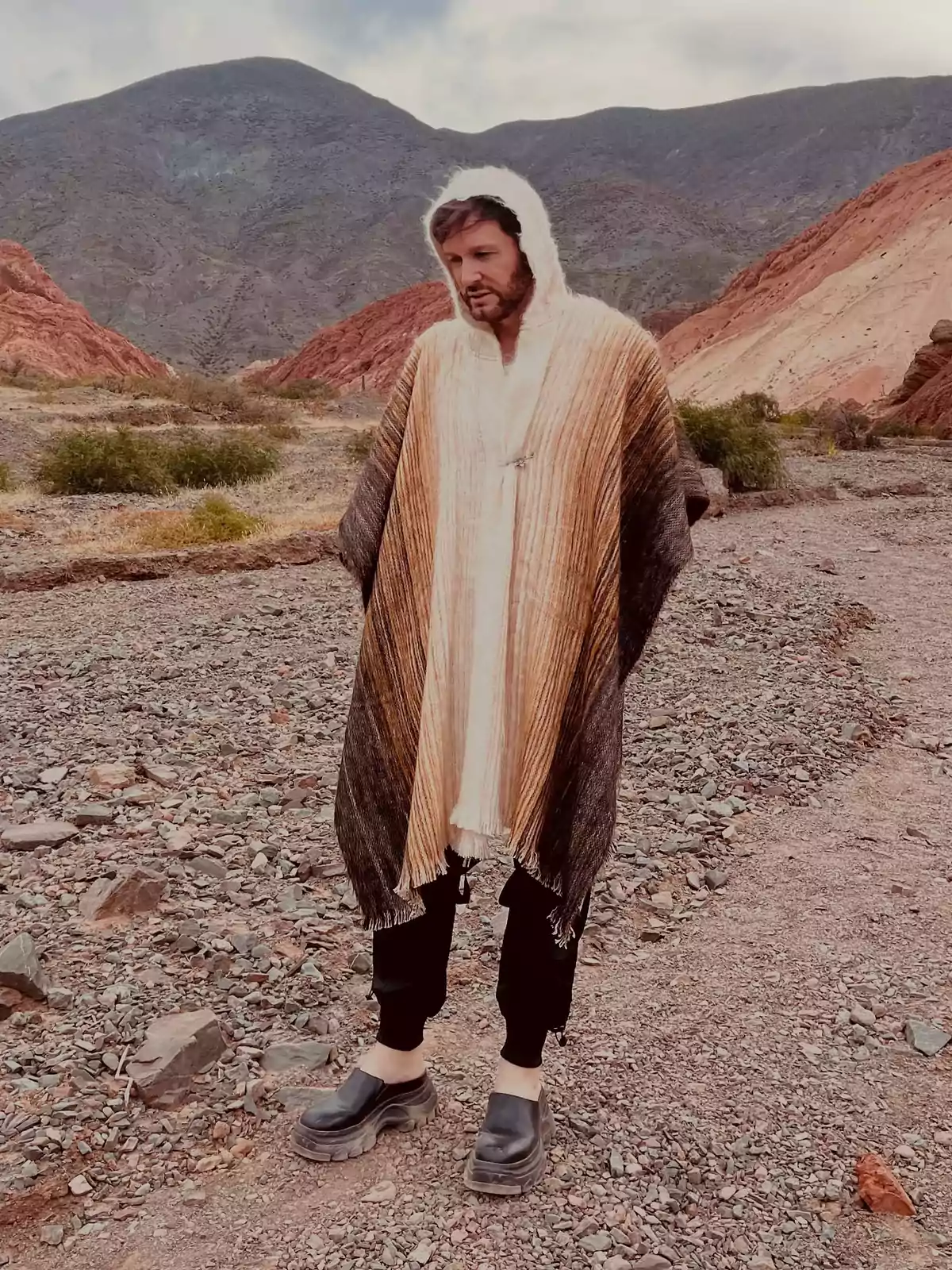 Person standing in an arid mountainous landscape wearing a brown and beige hooded poncho and dark pants Person standing in an arid mountainous landscape wearing a brown and beige hooded poncho and dark pants