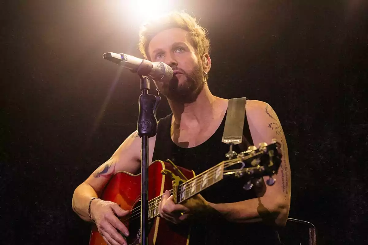 Bearded man with tattoos playing an acoustic guitar and singing into a microphone under a bright light on stage