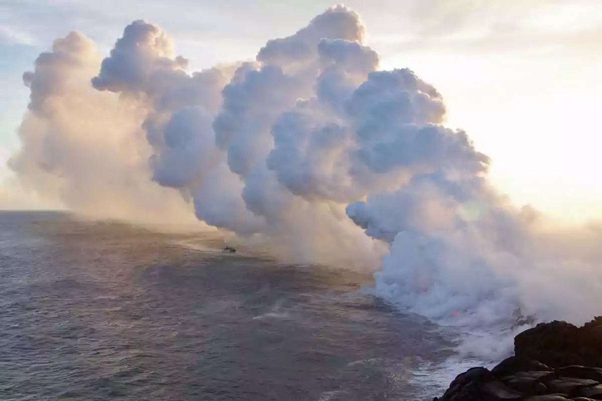 Grandes columnas de vapor se elevan donde la lava entra en contacto con el océano mientras un bote se acerca a la costa rocosa Grandes columnas de vapor se elevan donde la lava entra en contacto con el océano mientras un bote se acerca a la costa rocosa
