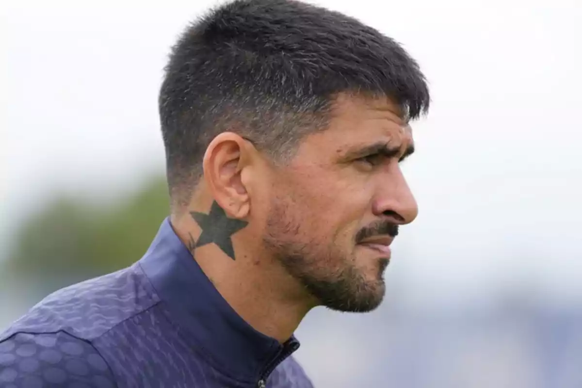 Hombre de perfil con barba corta y tatuaje de estrella negra en el cuello, vistiendo chaqueta deportiva azul Hombre de perfil con barba corta y tatuaje de estrella negra en el cuello, vistiendo chaqueta deportiva azul