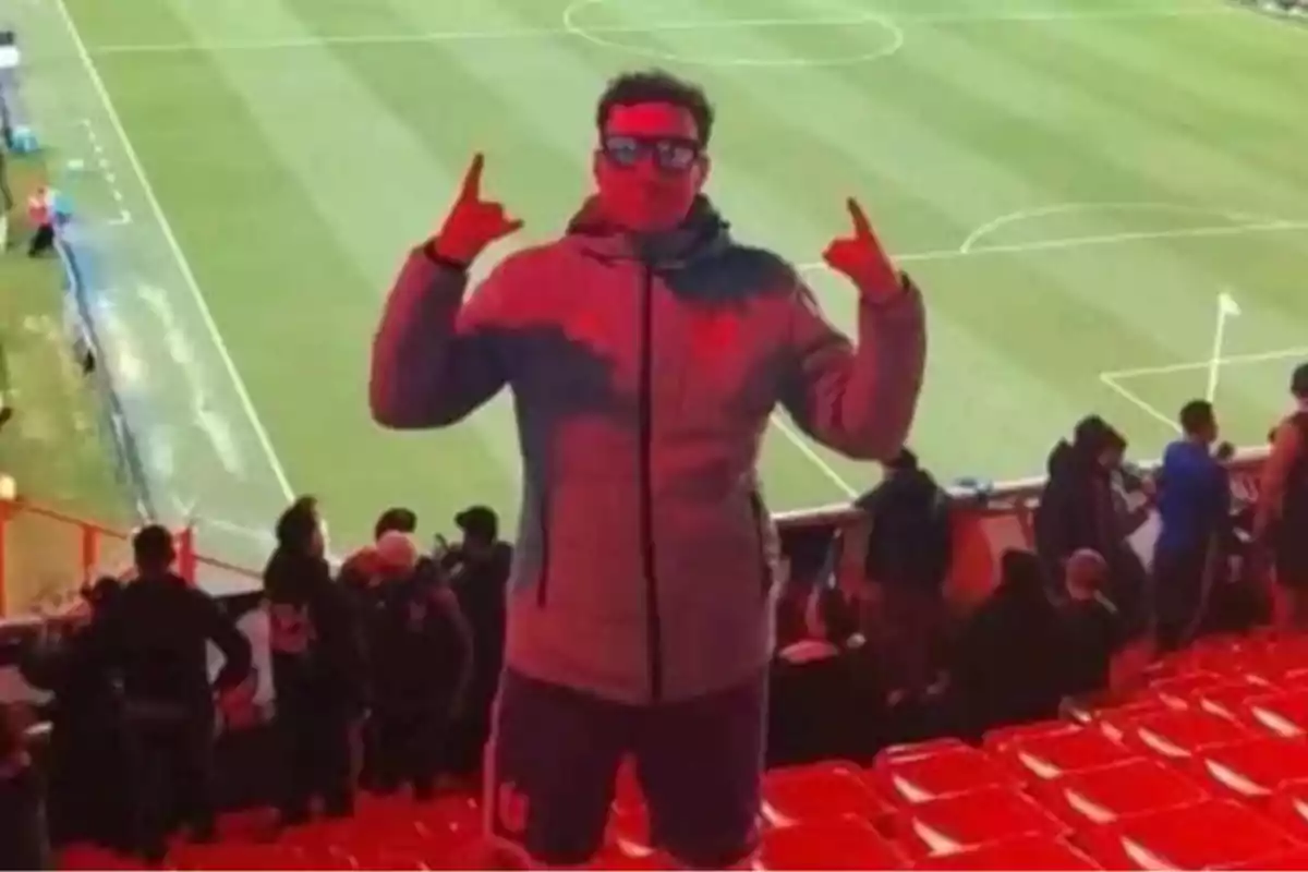 Persona con gafas y chaqueta posando en las gradas de un estadio de fútbol con el campo de juego al fondo y asientos rojos alrededor.