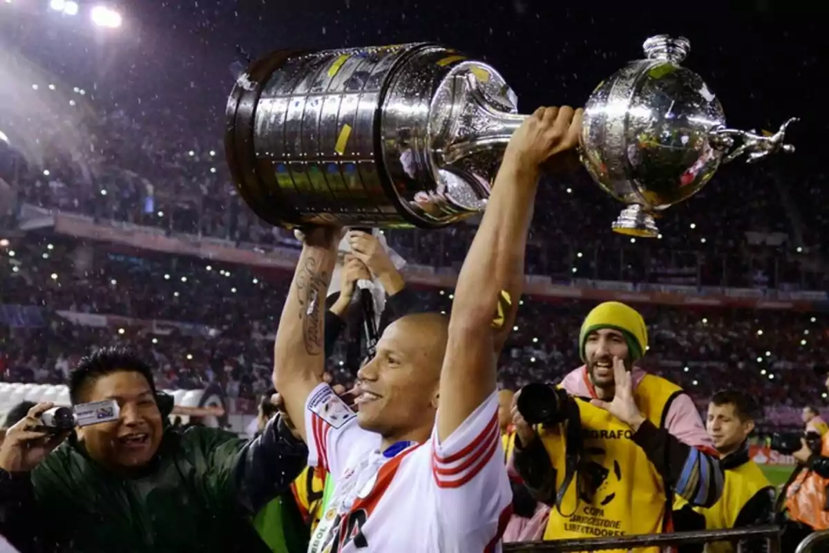 Soccer player celebrating with the Copa Libertadores trophy in a stadium full of fans and photographers