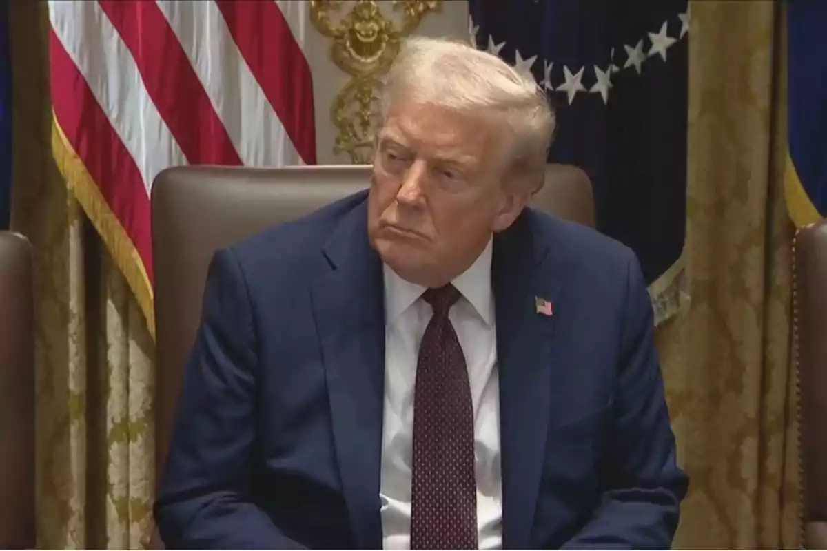 Blond-haired man in a navy blue suit and red tie sitting in a room with United States flags in the background