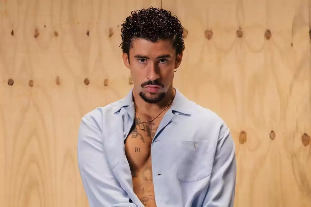 Hombre de cabello rizado y barba con camisa azul clara abierta mostrando tatuajes en el pecho posando frente a una pared de madera