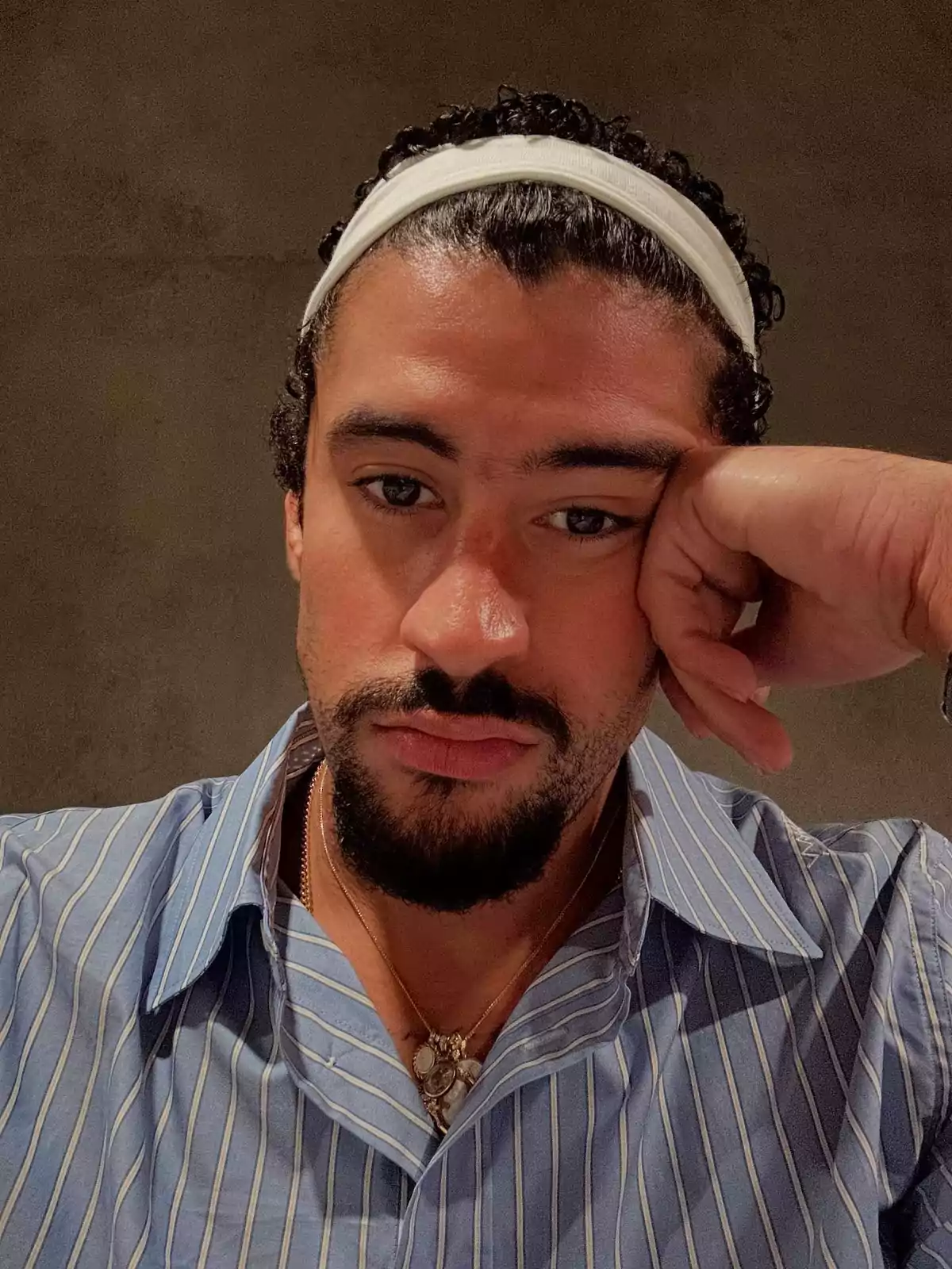 Hombre con barba y bigote usando una camisa de rayas azules y una banda blanca en la cabeza, apoyando su rostro en una mano y mirando a la cámara con expresión seria Hombre con barba y bigote usando una camisa de rayas azules y una banda blanca en la cabeza, apoyando su rostro en una mano y mirando a la cámara con expresión seria