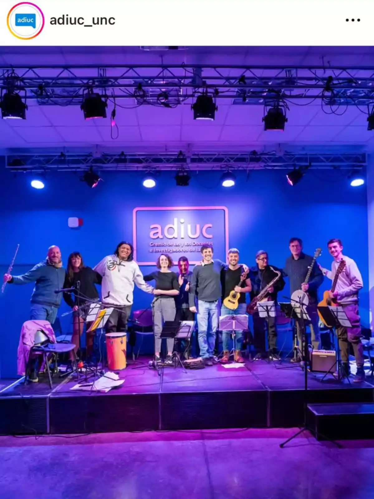 A group of people smiles on a stage lit with violet and blue lights; some are holding musical instruments like a guitar, saxophone, and drum, and behind them there is a sign that says adiuc. A group of people smiles on a stage lit with violet and blue lights; some are holding musical instruments like a guitar, saxophone, and drum, and behind them there is a sign that says adiuc.