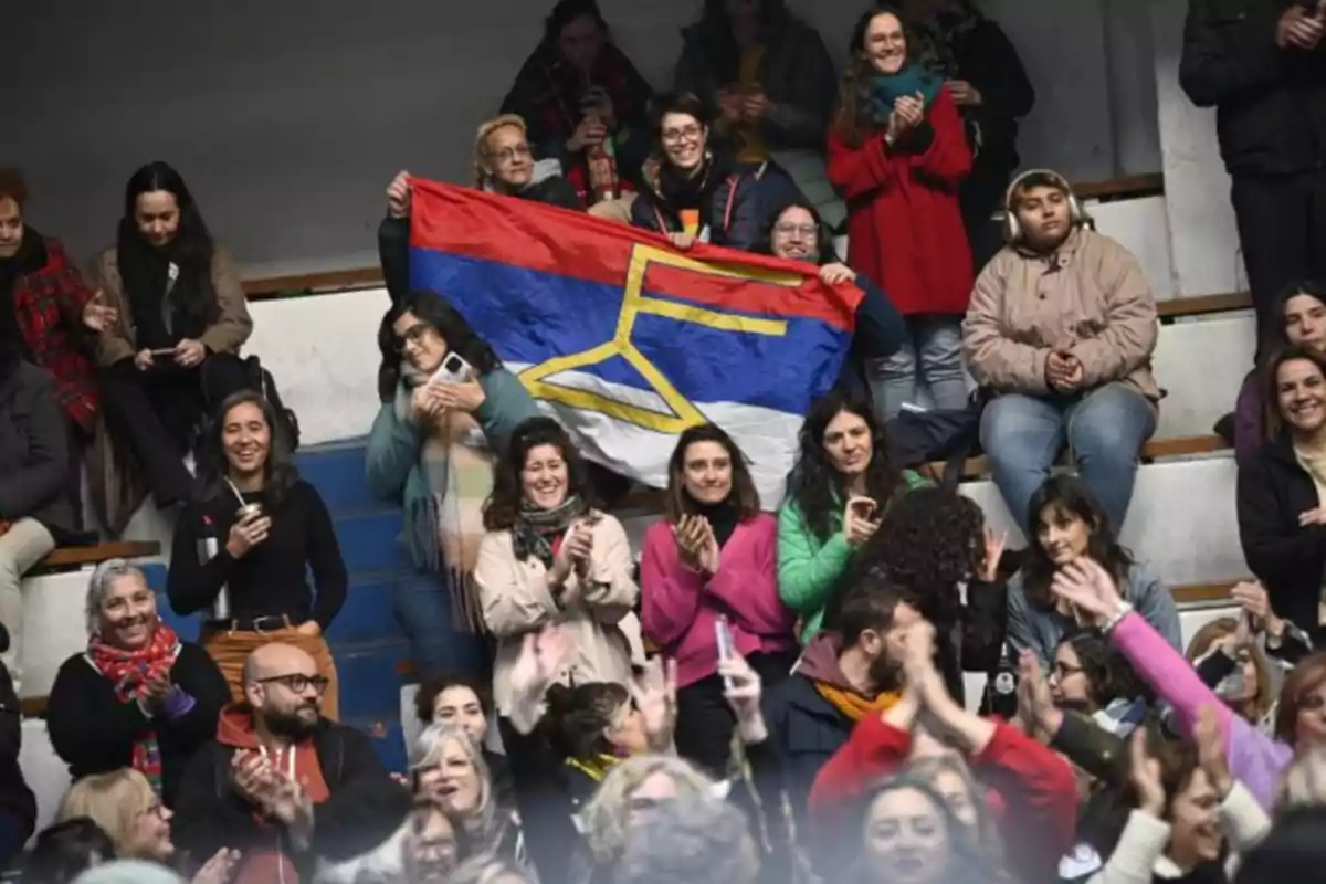 People sitting and standing on bleachers, some applauding and others smiling, while a group holds a tricolor flag with a yellow symbol in the center. People sitting and standing on bleachers, some applauding and others smiling, while a group holds a tricolor flag with a yellow symbol in the center.