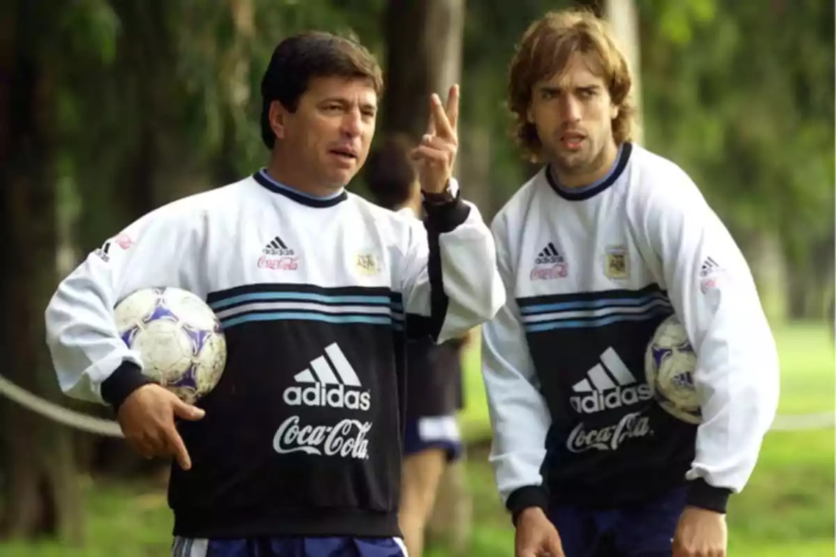 Two men wearing Argentina national soccer team training gear hold balls on a field surrounded by trees. Two men wearing Argentina national soccer team training gear hold balls on a field surrounded by trees.