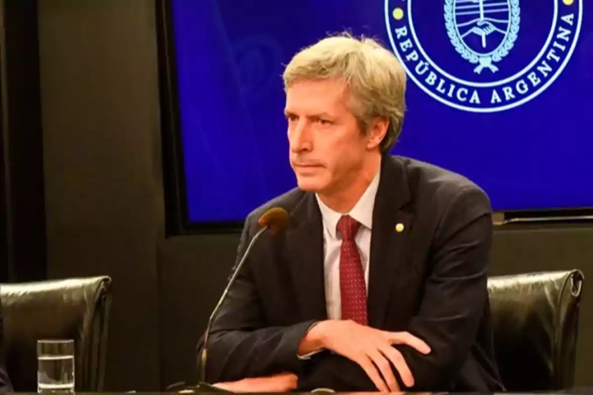 Gray-haired man in a dark suit and red tie sitting in front of a microphone at a press conference with the coat of arms of the Argentine Republic in the background Gray-haired man in a dark suit and red tie sitting in front of a microphone at a press conference with the coat of arms of the Argentine Republic in the background