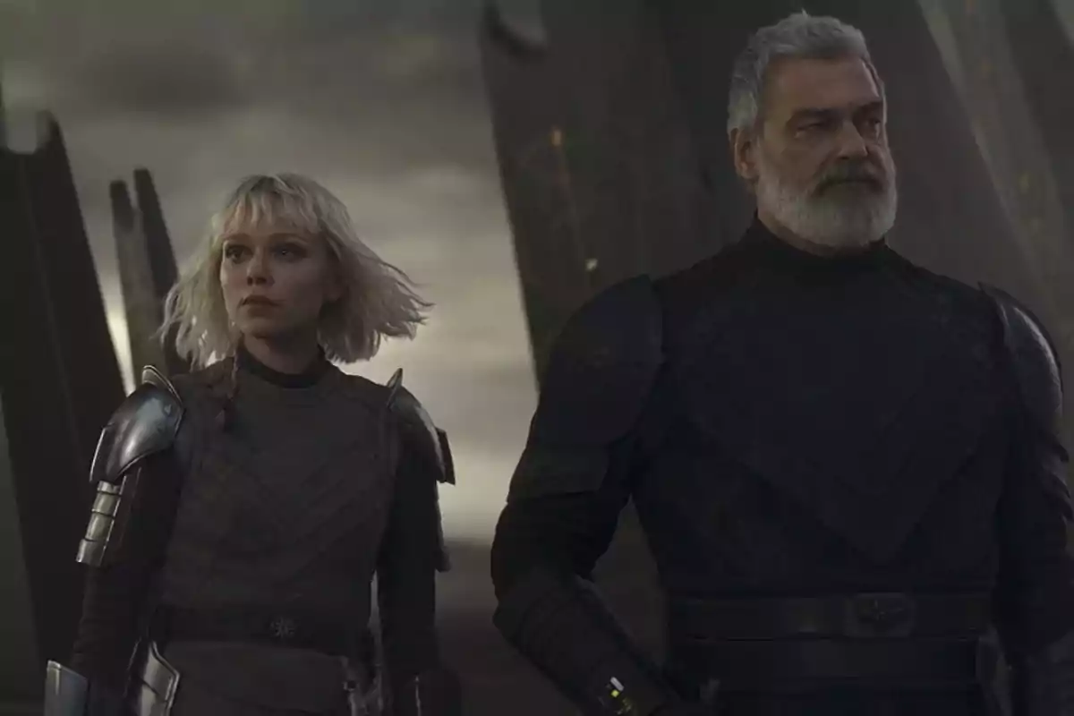 Two people in dark armor look toward the horizon in a somber, futuristic setting. Two people in dark armor look toward the horizon in a somber, futuristic setting.