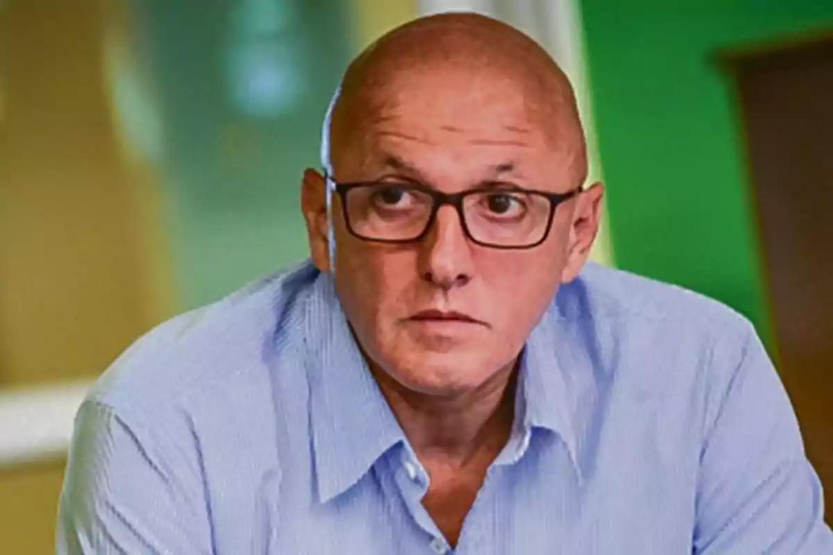 Hombre calvo con gafas y camisa azul claro mirando hacia el frente en un entorno interior desenfocado
