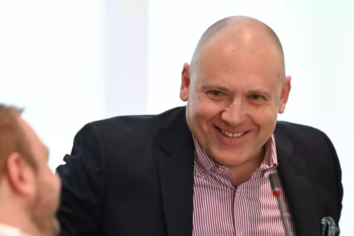 Bald man in a striped jersey and dark jacket smiling while talking to another person