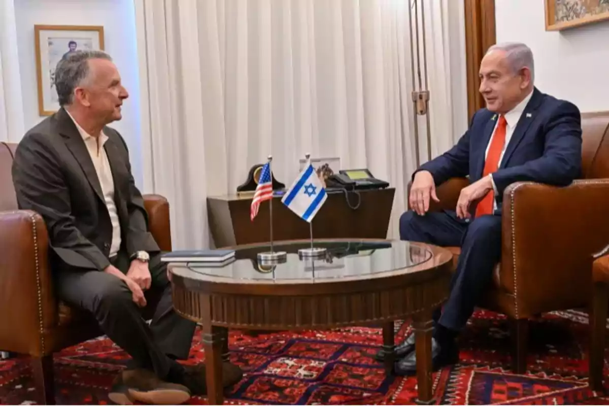 Two men in suits are sitting in brown leather armchairs, talking across a glass table with small flags of the United States and Israel. Two men in suits are sitting in brown leather armchairs, talking across a glass table with small flags of the United States and Israel.