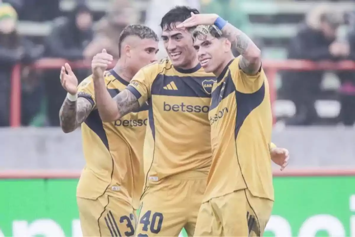Three players in yellow uniforms celebrate a goal in a stadium under the rain.