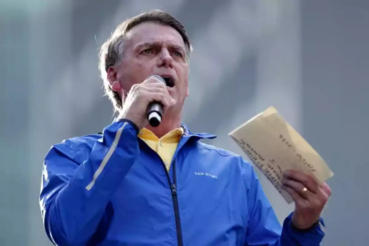 Man in a blue jacket speaking into a microphone and holding papers in his hand