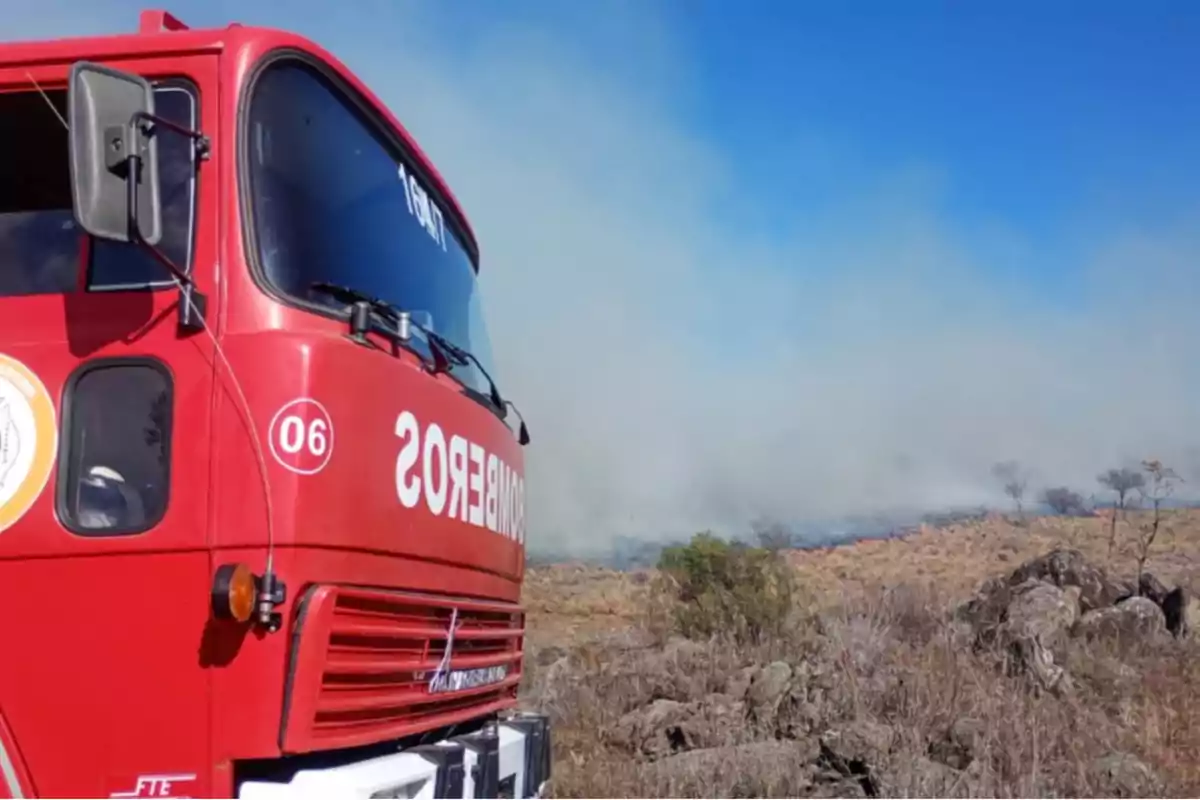 Camión de bomberos rojo en primer plano mientras al fondo se observa un incendio forestal con humo y vegetación seca Camión de bomberos rojo en primer plano mientras al fondo se observa un incendio forestal con humo y vegetación seca