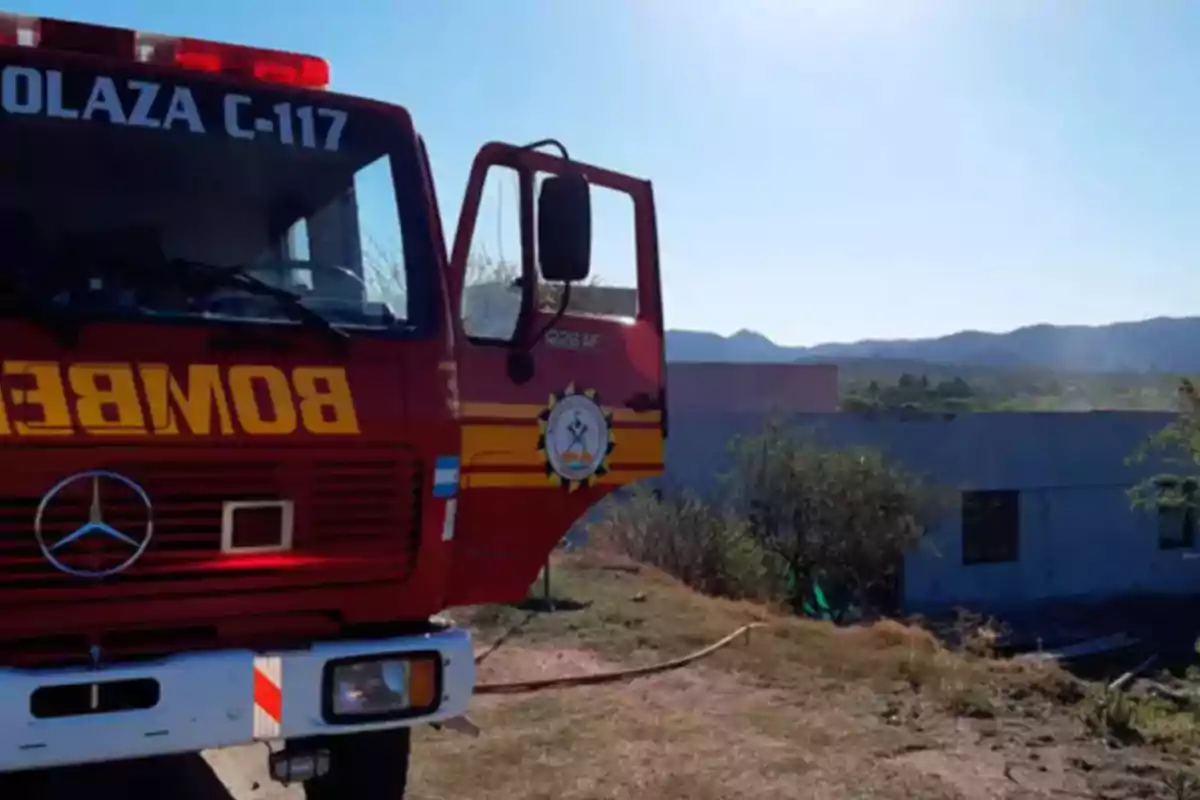 Camión de bomberos rojo estacionado en un terreno con vegetación y una casa al fondo en un día soleado