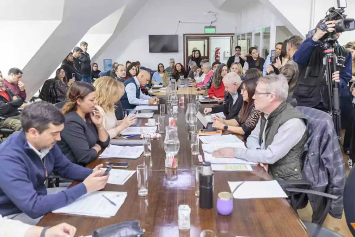 Personas reunidas alrededor de una mesa larga en una sala de reuniones, algunas usando computadoras y teléfonos, mientras otras toman notas y conversan, con cámaras y periodistas al fondo.
