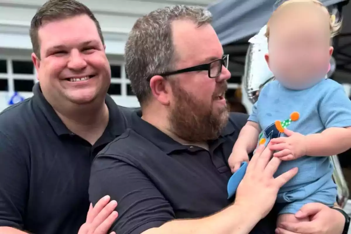 Three people pose together outdoors, two adults are smiling, and one of them is holding a baby with a blurred face.