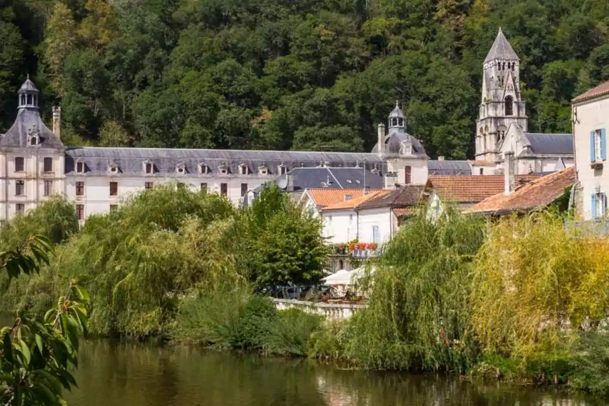 Edificio histórico con torre y tejados grises rodeado de árboles y vegetación junto a un río Edificio histórico con torre y tejados grises rodeado de árboles y vegetación junto a un río