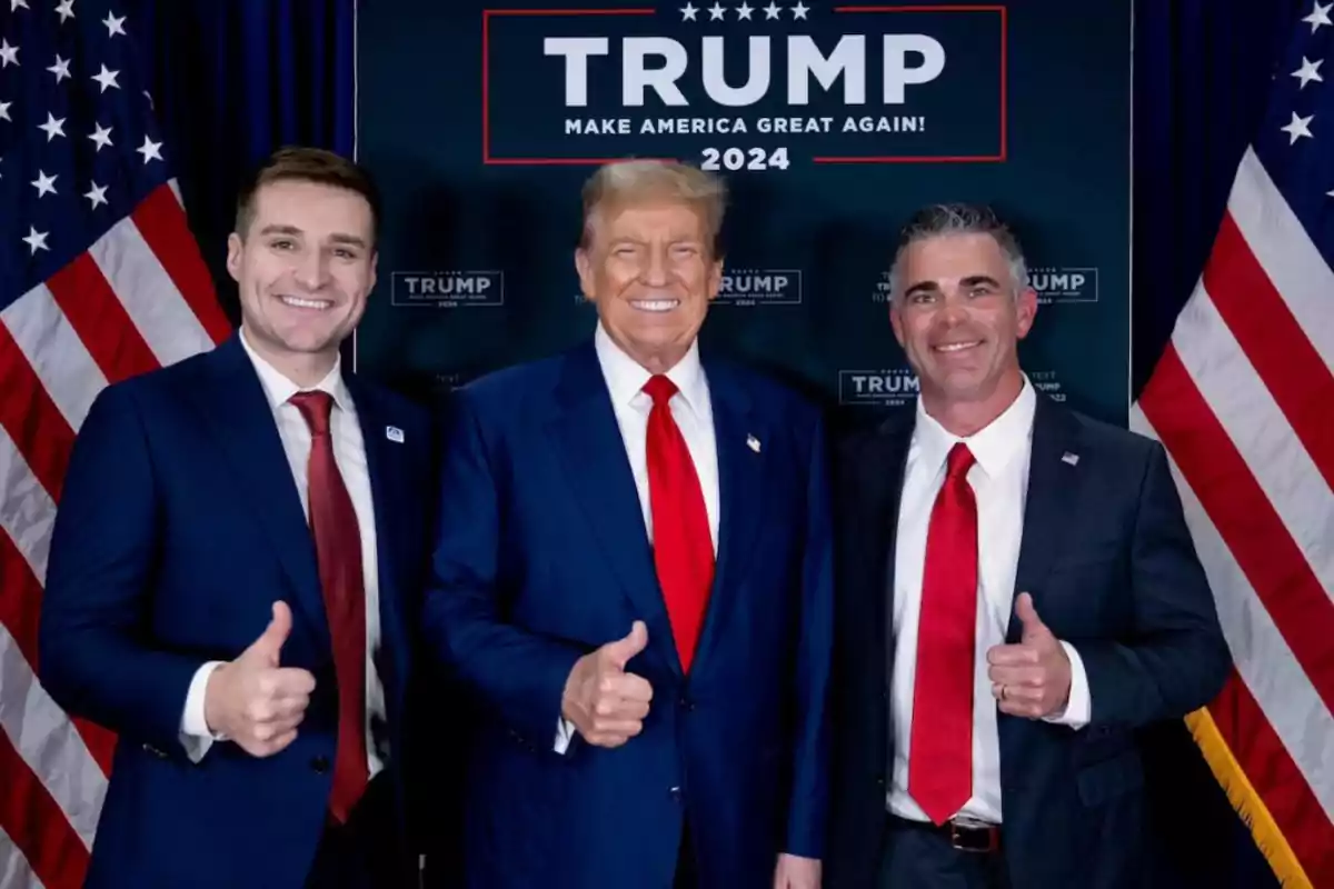 Three men in suits and red ties pose smiling and giving a thumbs up in front of a background with American flags and a Trump 2024 campaign sign. Three men in suits and red ties pose smiling and giving a thumbs up in front of a background with American flags and a Trump 2024 campaign sign.