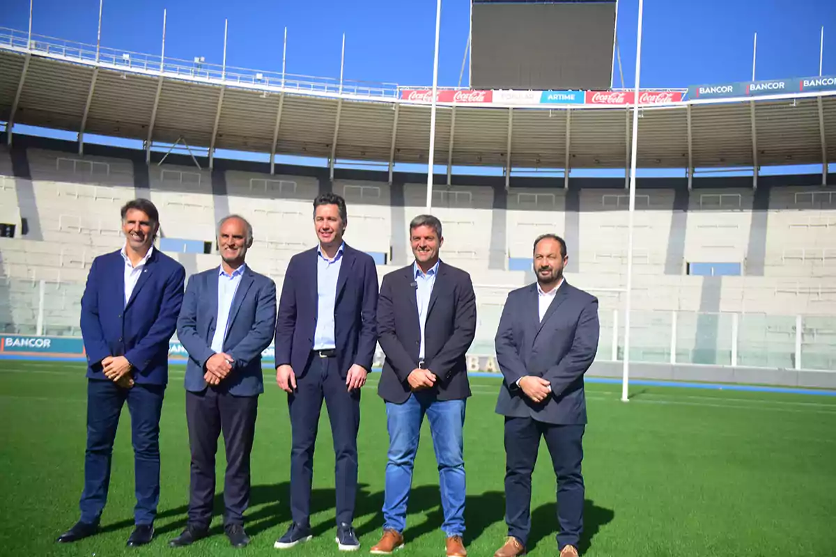 Cinco hombres de traje posan sobre el césped de un estadio de fútbol vacío en un día soleado