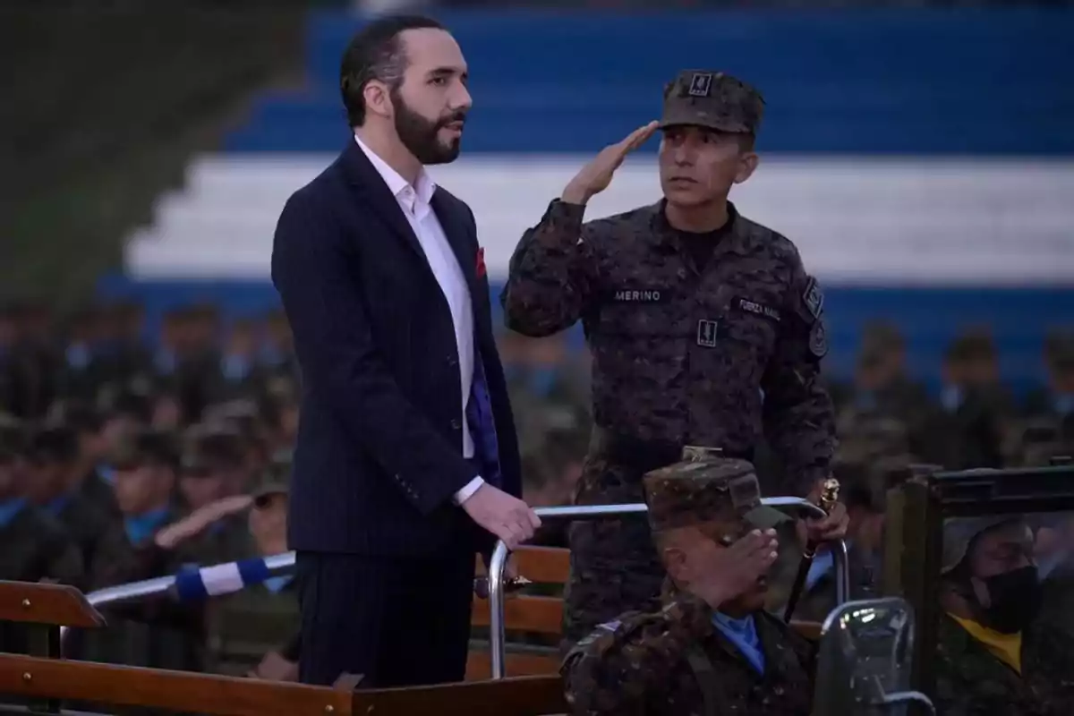 A man in a dark suit is standing next to a saluting soldier, both on a vehicle at an event with soldiers in the background.