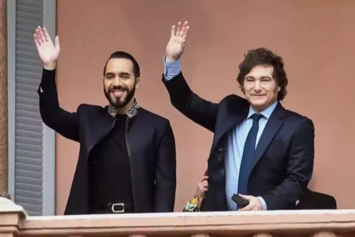 Two men standing on a balcony wave and smile, one wearing a dark suit and the other dressed in black with details on the collar.