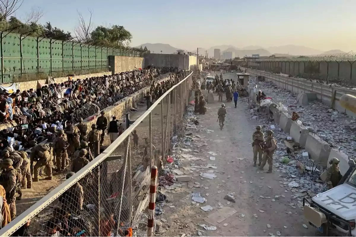 Soldiers patrol a fenced corridor while a crowd of people waits sitting next to a fence in an area filled with trash and debris. Soldiers patrol a fenced corridor while a crowd of people waits sitting next to a fence in an area filled with trash and debris.