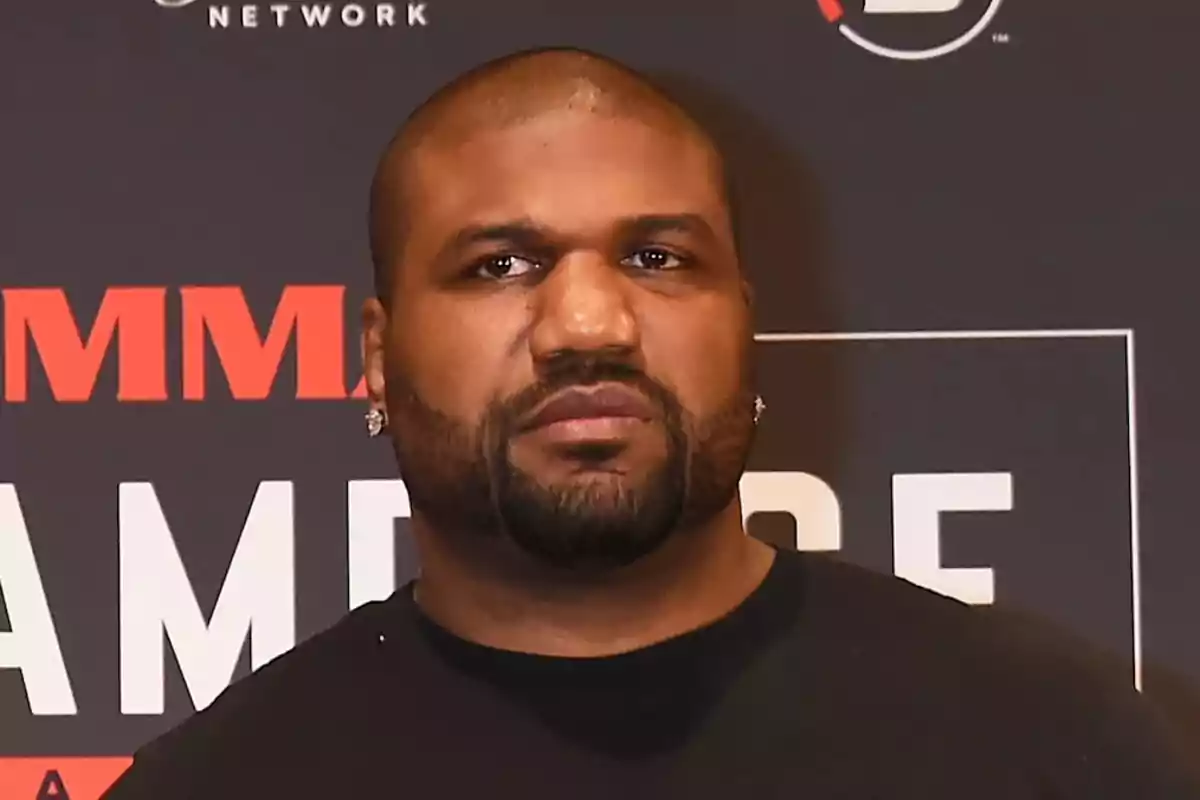 Hombre de expresión seria con barba y camiseta negra posando frente a un fondo con letras y logotipos Hombre de expresión seria con barba y camiseta negra posando frente a un fondo con letras y logotipos