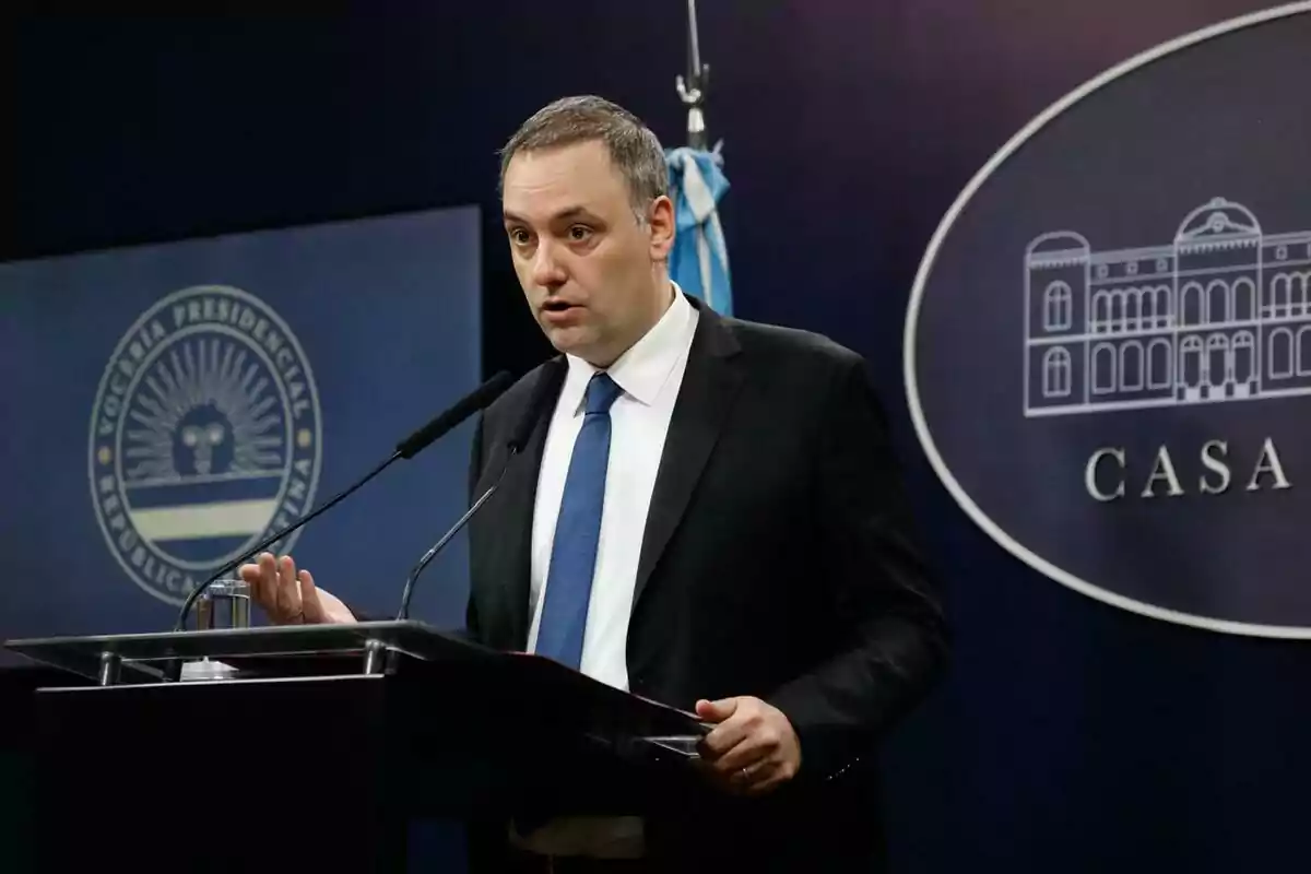 A man in a blue suit and tie speaks at a press conference in front of microphones with flags and official emblems in the background. A man in a blue suit and tie speaks at a press conference in front of microphones with flags and official emblems in the background.