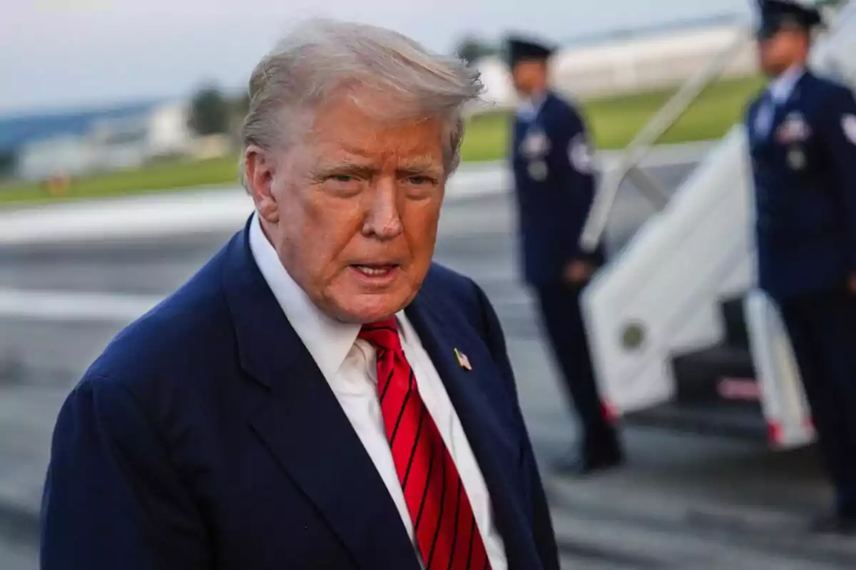 Hombre de cabello rubio con traje azul marino y corbata roja en un aeropuerto con personal militar al fondo Hombre de cabello rubio con traje azul marino y corbata roja en un aeropuerto con personal militar al fondo
