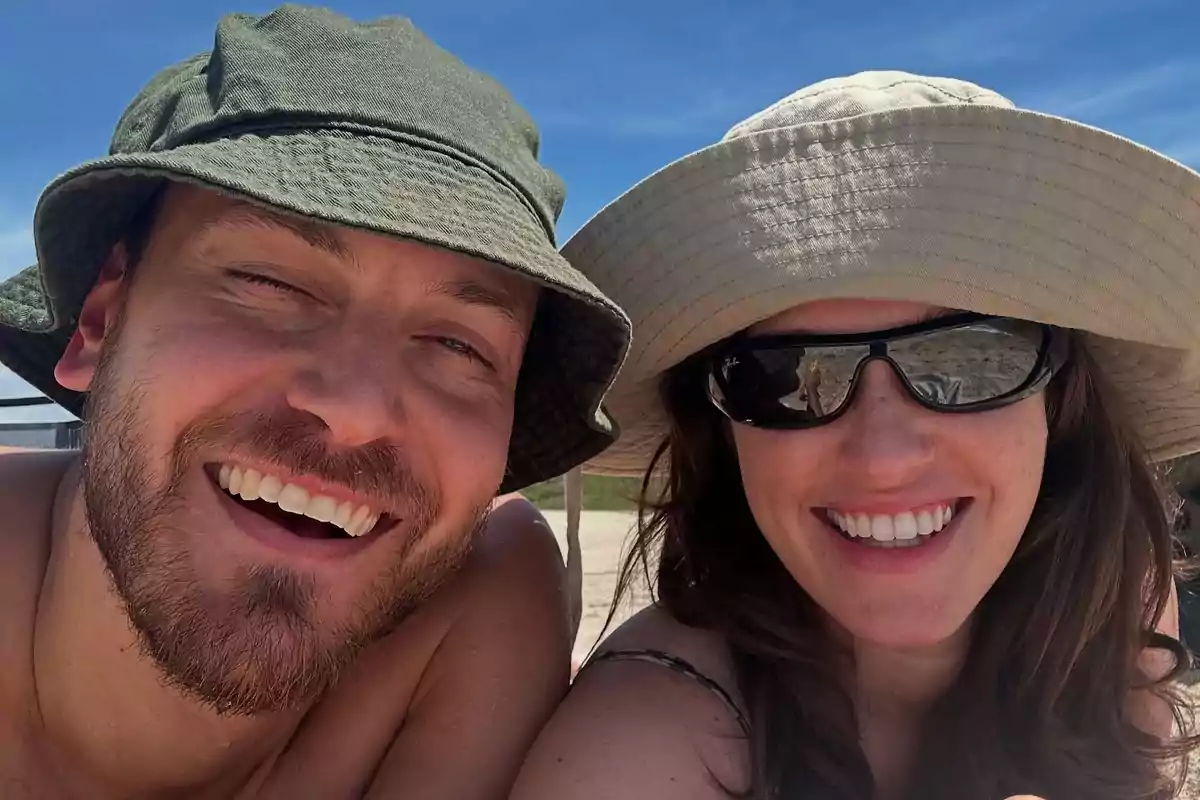 Pareja sonriendo en la playa usando sombreros y gafas de sol bajo un cielo azul Pareja sonriendo en la playa usando sombreros y gafas de sol bajo un cielo azul