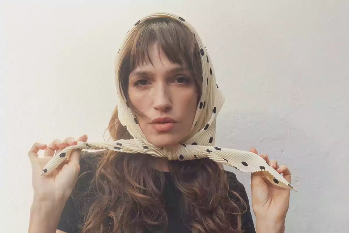 Woman with long brown hair wearing a beige scarf with black polka dots on her head in front of a white wall